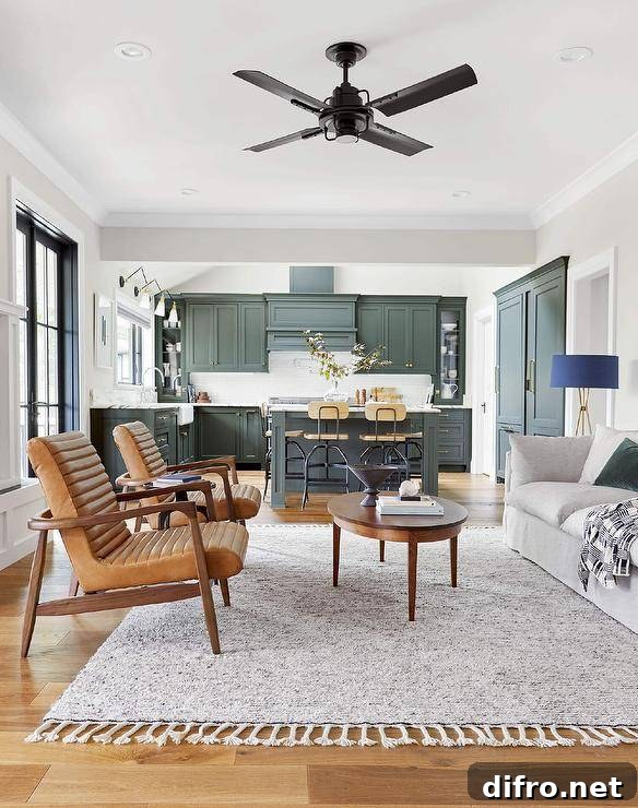 A black ceiling fan cools an open living room featuring an oval wooden coffee table placed on a gray fringe rug and flanked by a pebble gray sofa topped with an emerald green velvet pillow and mid-century modern leather and wooden chairs.