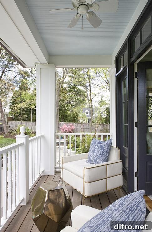 Gorgeous covered porch features a blue beadboard ceiling accented with a ceiling fan over ivory upholstered chairs with brass trim adorned with blue medallion pillows alongside a brass geometric stool table.