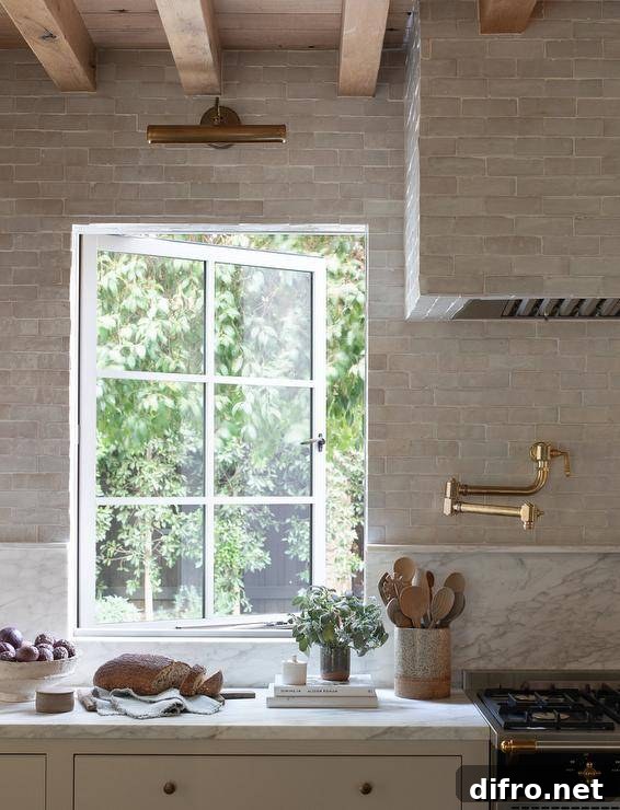 A brass picture light is fixed to gray glazed backsplash tiles over a casement window partially framed by a marble backsplash. A gray glazed tiled hood is mounted over a brass pot filler.