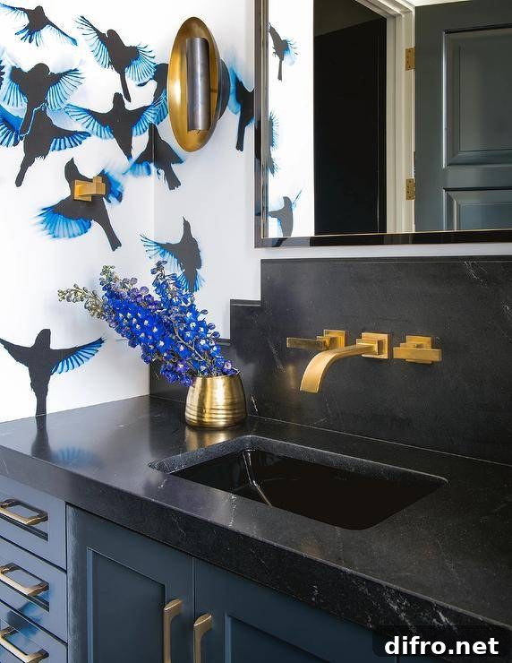 Chic black and blue bathroom features walls covered in blue and black wallpaper and a gold and black mirror hung above an aged brass faucet mounted to a honed black marble backsplash over a blue washstand.