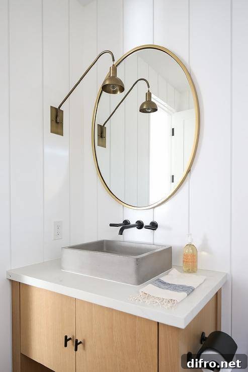 A gold vanity mirror is hung from a white vertical plank trim and lit by a brass swing arm sconce. Beneath the mirror, an oil rubbed bronze wall mount faucet is fitted above a concrete vessel sink mounted to a bronze sink vanity.