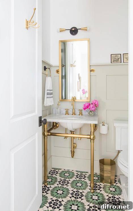 A marble and brass washstand is fixed against a light taupe wainscot wall to green and black cement mosaic floor tiles. Above the sink vanity, a tall brass pivot mirror is lit by a brass and gold 2-light sconce.