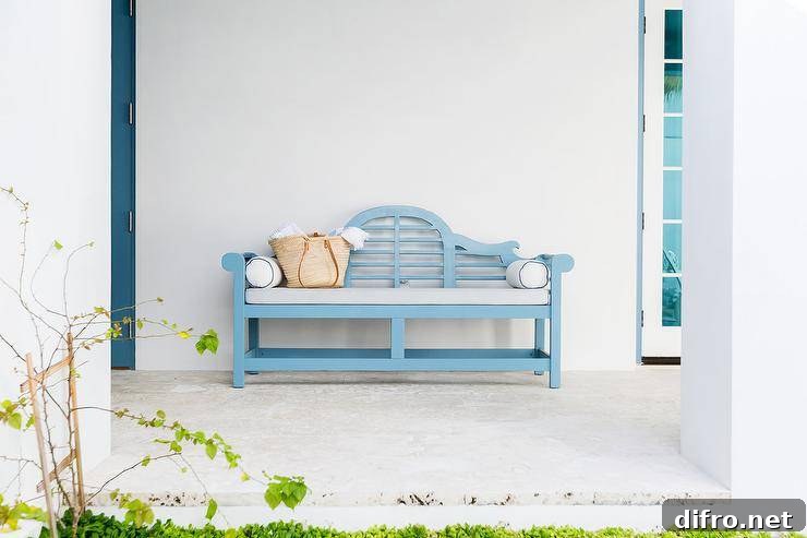 Outdoor cottage patio charmingly designed with a blue wooden bench with a light gray cushion. White exterior walls and lush greenery offer a streamlined template for added decor.