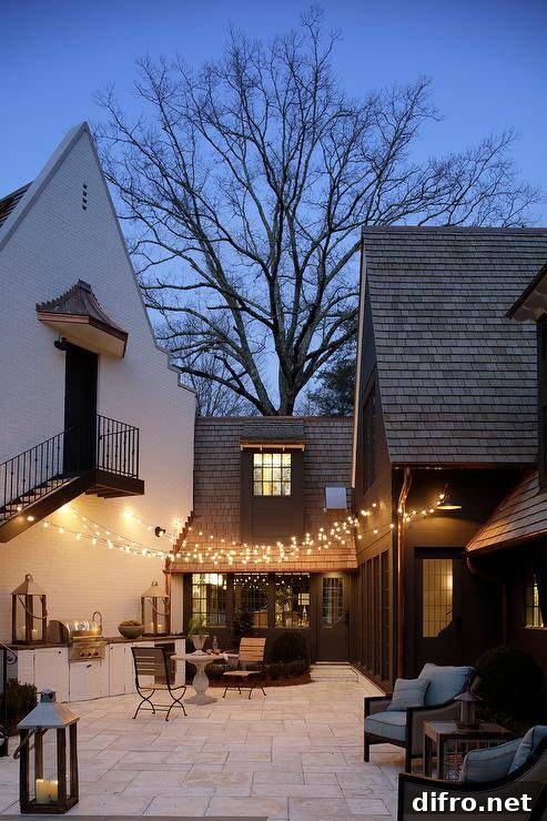 String lights and large outdoor candle lit lanterns illuminate two wood and iron cafe chairs sitting in front of a round marble cafe table.