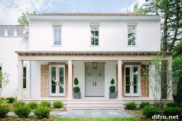 Stunning white brick cottage home with soft gray French front doors, vintage wood shutters and a covered porch with decorative plants.