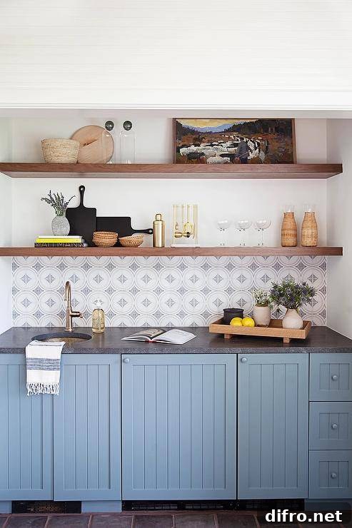Brown wooden floating shelves stacked above white and gray mosaic tiles above blue cabinets with a round hammered brass sink.