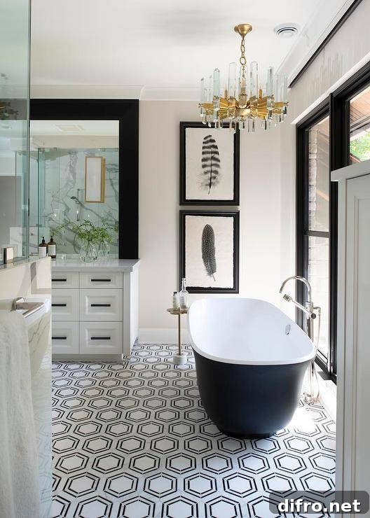 A lucite and brass chandelier lights a black and white cast iron bathtub placed on black and white hexagon floor tiles and paired with a floor mount chrome tub filler. The tub filler is mounted in front of black framed windows, while stacked feather art in black frames is hung behind the bathtub.