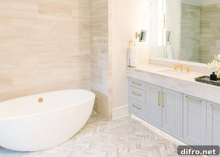 Stunning master bathroom with a light gray washstand and light gray marble countertop displaying a white mirror and brass hardware. under cabinet lighting upgrades this design with a luxurious appeal highlighting white and gray herringbone floor tiles. An oval freestanding tub against a light gray staggered tiled wall invites a spa like look to the bathroom appeal.