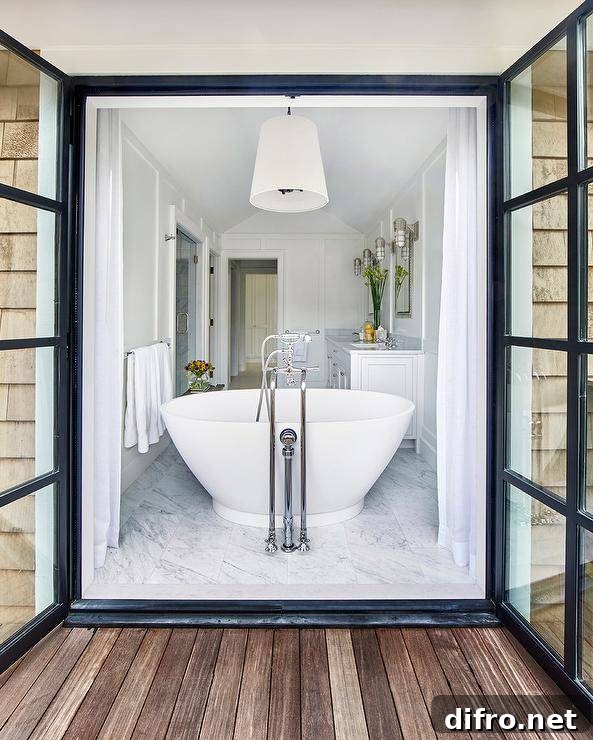 White cottage bathroom features a white tapered drum pendant light which hangs over a freestanding bathtub placed in front of steel doors dressed in white curtains which open to the deck.