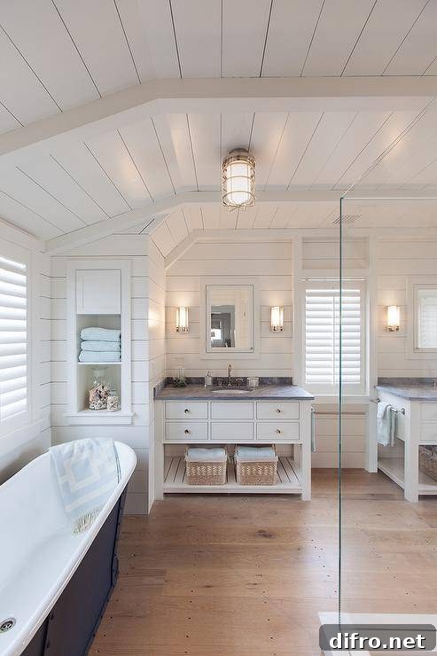 Fantastic bathroom features a shiplap ceiling and walls lined with a freestanding tub placed under windows covered in plantation shutters atop sawn white oak wood floors. Master bathroom boasts separate white washstands with shelf topped with honed Bardiglio marble under inset white framed medicine cabinets.
