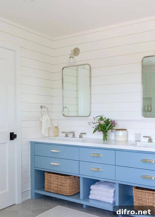 White and blue cottage bathroom boasts a blue dual washstand placed on charcoal gray herringbone floor tiles and fitted with shelves holding woven bins. The washstand is accented with polished nickel cup pulls and polished nickel faucets fixed beneath curved nickel mirrors. The mirror are hung over white staggered backsplash tiles and mounted to white shiplap under glass and nickel conical sconces.