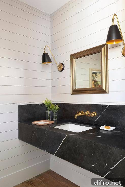 Fixed to shiplap trim, gold and black sconces flank a square gold beveled mirror is hung over a honed black marble floating sink vanity finished with a brass faucet kit fixed to a black marble backsplash.