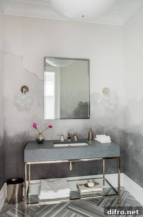 A nickel framed mirror hangs from a wall covered in a white and gray mural and between white glass globe sconces lighting gray marble herringbone floor tiles. Beneath the mirror, a stone and nickel sink vanity is fitted with a glass shelf and a polished nickel faucet.