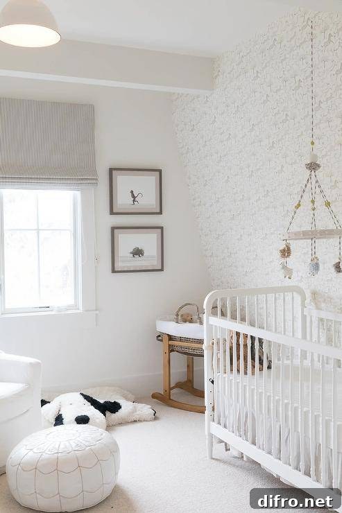 Lovely monochromatic nursery boasts a sheep mobile hung in front of an accent wall covered in white and gray horse print wallpaper and over a white crib. The crib is accented with a white ruffled skirt and placed on ivory carpeting. Stacked art hangs beside a window covered in a stripe gray roman shade.