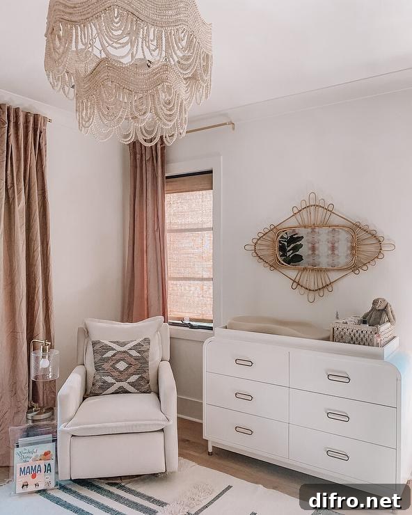 Welcoming nursery is illuminated by a beaded chandelier and features a modern white curved dresser topped with a changing pad and positioned under a rattan mirror. An acrylic accent table accented with a nickel and glass lamp is placed beside an off-white glider, while windows are covered in brown linen curtains.