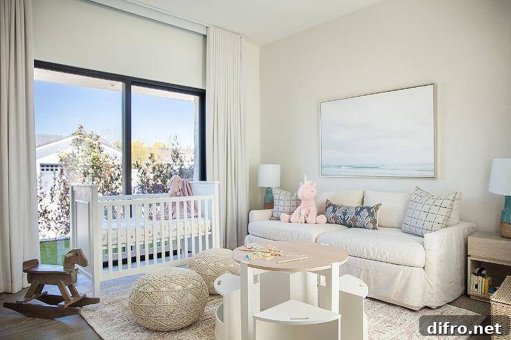 Welcoming nursery is styled with macrame poufs placed on a gray rug at a round two-tone play table with white chairs. n ocean art piece hangs over an off-white couch topped with gray and purple pillows lit by turquoise blue cork lamps. A white crib sits in front of sliding glass doors covered in white curtains.