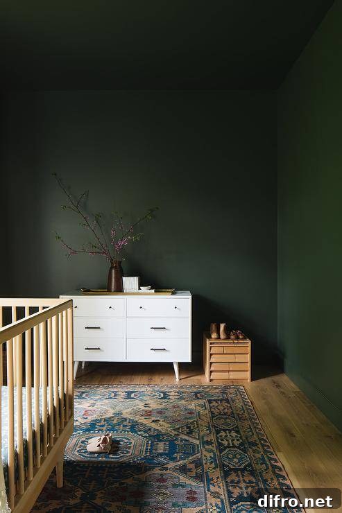Forest green nursery walls and ceiling complement a white mid-century modern dresser placed behind a pink and blue vintage rug positioned beneath a blond wood crib.