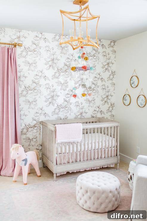 A brass lantern illuminates a stunning pink, white, and gray nursery. Pink curtains hang from a brass rod beside a gray French crib placed in front of a black and white floral wallpapered wall and beneath a pom pom mobile.