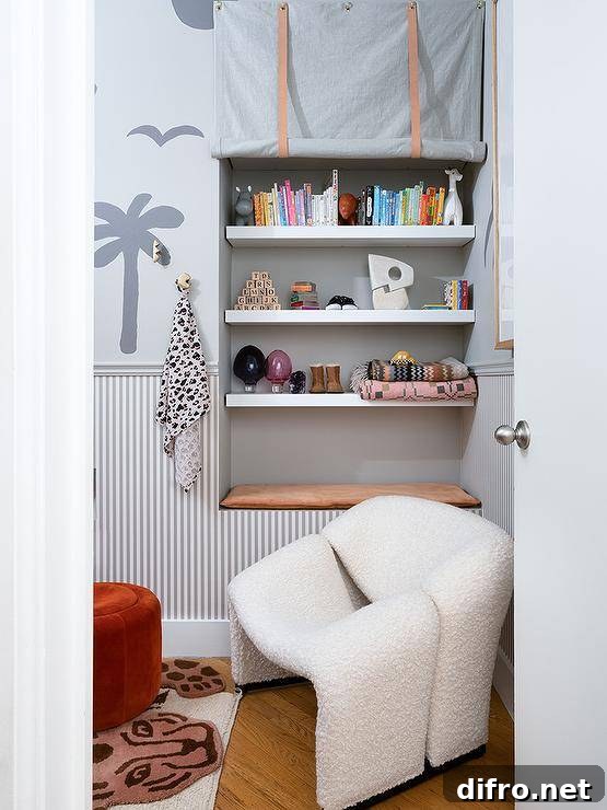 A white boucle chair sits in a modern nursery next to an alcove fitted with a tan cushion located beneath white shelves covered in a gray rolling curtain. A gray chair rail separates gray pinstripe wallpaper and gray gray patterned wallpaper.