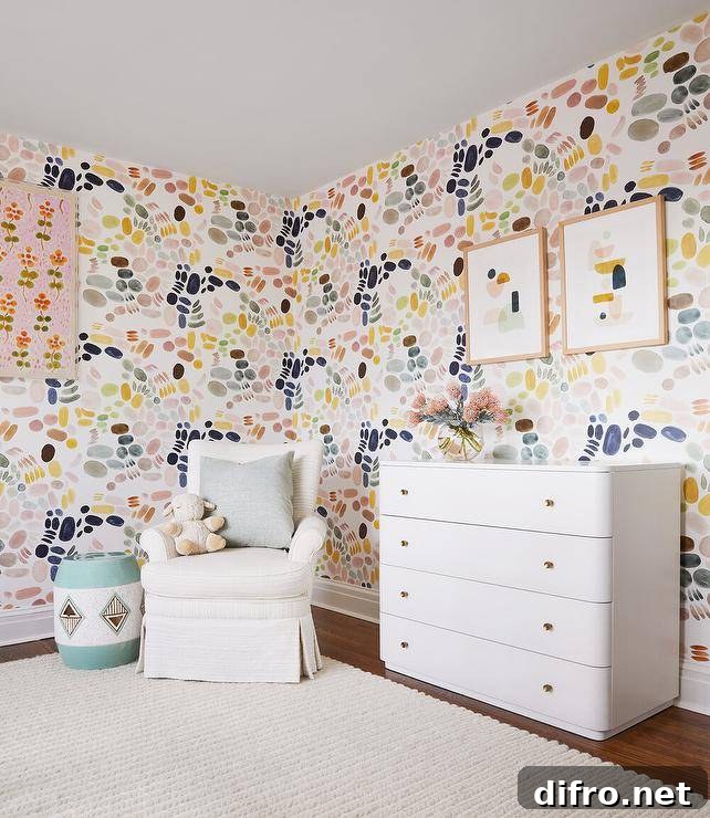 Girl's nursery features pink blue green yellow and gray wallpaper, a white nursery dresser with brass knobs and an ivory roll arm glider over a cream rug.