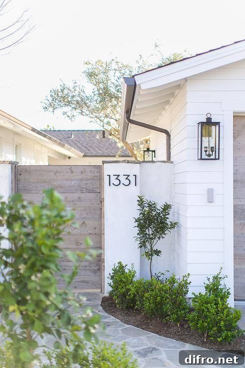 Exterior home features modern steel house numbers displayed on a white finished wooden house gate. Winding stone pavers sets a surface toward the wooden gate into a finished backyard.
