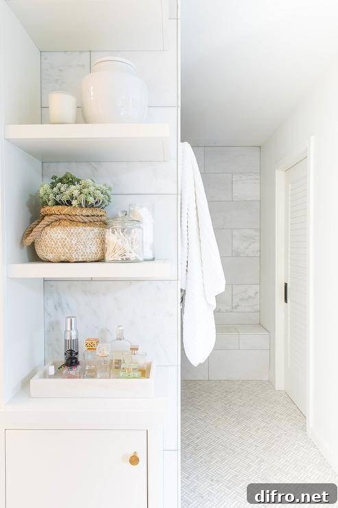 Stacked white floating bathroom shelves are mounted against staggered marble tiles over a white flat front cabinet adorned with a perfume bottle tray.