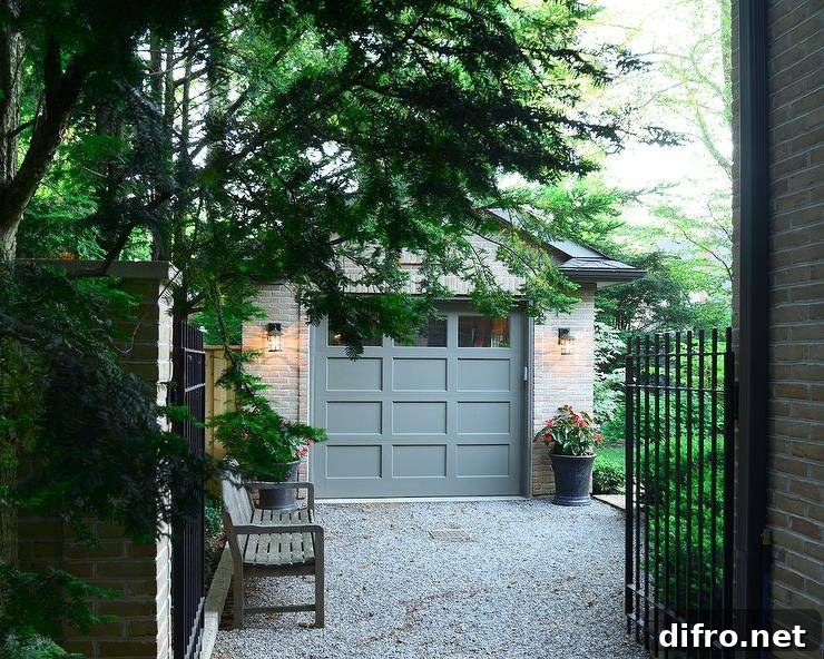 Iron gates open to a gravel driveway leading past a wooden bench to a brick garage finished with a gray multi-panel garage door flanked by carriage lanterns fixed over black planters.