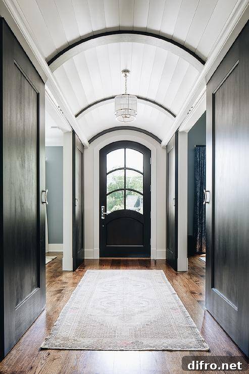 Long hallway with a plank barrel ceiling elegantly displaying a cascading chandelier over a light gray vintage rug layered on oak floors. A black arch front door brings a unique look contrary to a dull standard door.