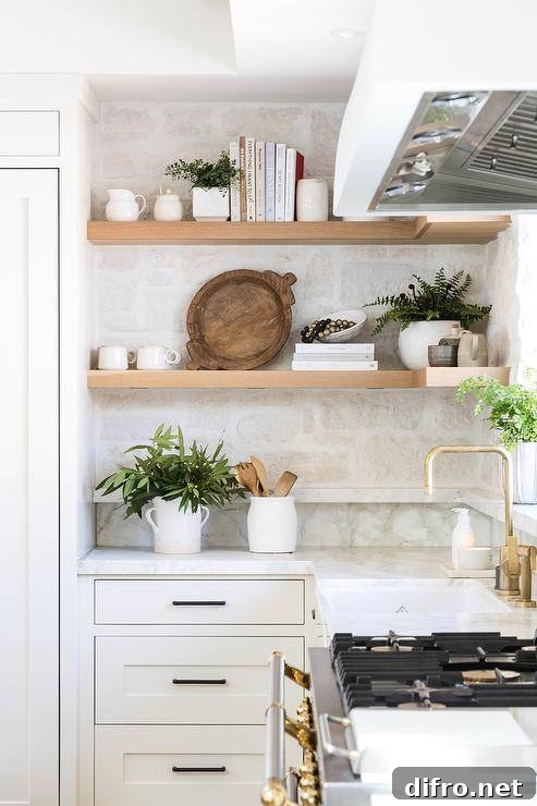 Kitchen features blond floating shelves on a rustic stone backsplash and white cabinetry accented with bronze pulls.