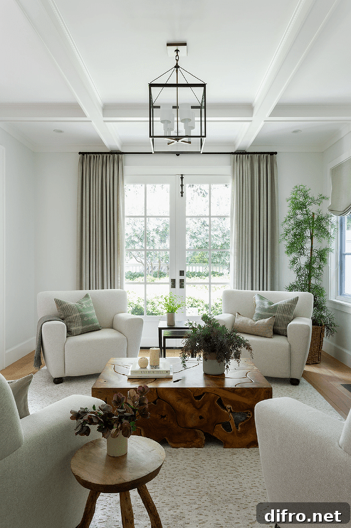 Living room features sage green pillows on light gray accent chairs, a burl wood coffee table atop a light gray rug, a round wooden stool as accent table and a tall potted plant.