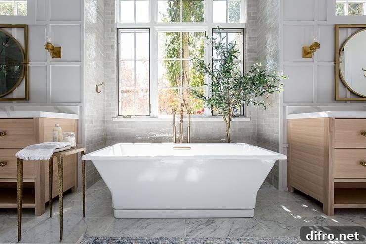 Brushed gold floor mount tub filler paired with a rectangular freestanding tub in a gray tiled bathtub nook. A corner house plant accents the nook.
