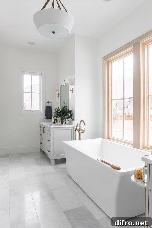 A rectangular freestanding bathtub with a polished nickel floor mount tub filler sits beneath a window on and between gray his and hers washstands.