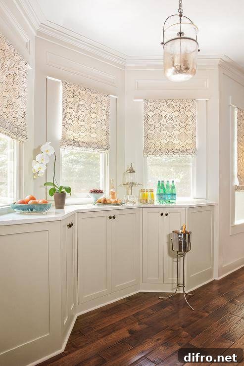 Kitchen bay window with tan shades features ivory built in cabinets accented with oval bronze knobs and white and silver marble countertops and dark wooden floors, illuminated by a mercury glass lantern.