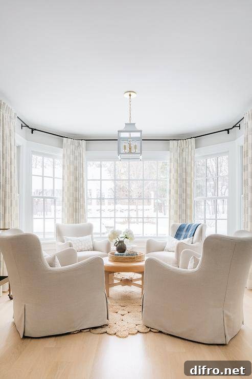 A white pagoda lantern hangs over a round brown leather ottoman placed on a crochet rug ad surrounded by white wingback lounge chairs. The chairs sit in front of bay windows dressed in ivory and black curtains.