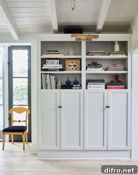 An antique brass picture light is fixed beneath a white plank ceiling and over white built-in bookshelves, styled and fitted over white shaker built-in cabinets donning oval knobs.