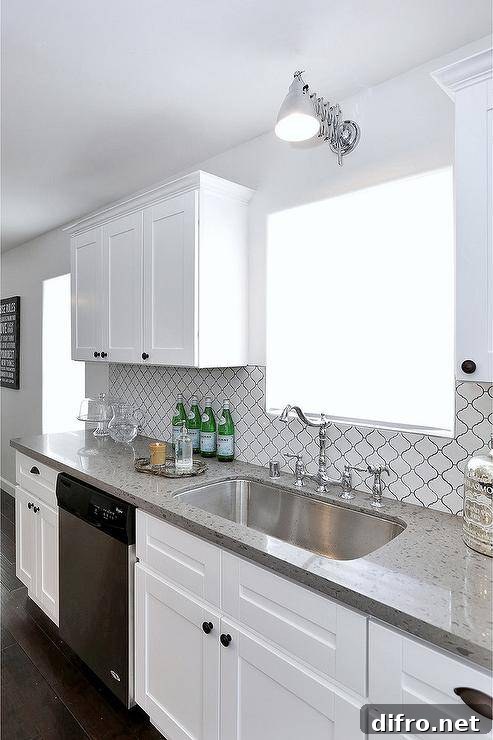 Well appointed white kitchen features a stainless steel dishwasher fixed between white shaker cabinets accented with oil rubbed bronze knobs and a gray quartz countertop.