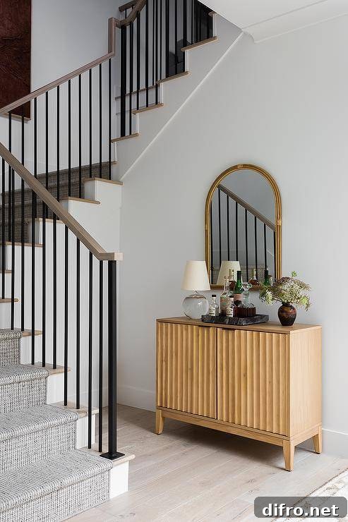 A tan wood bar cabinet accented with a crystal lamp placed beside a staircase beneath an arch bamboo mirror.