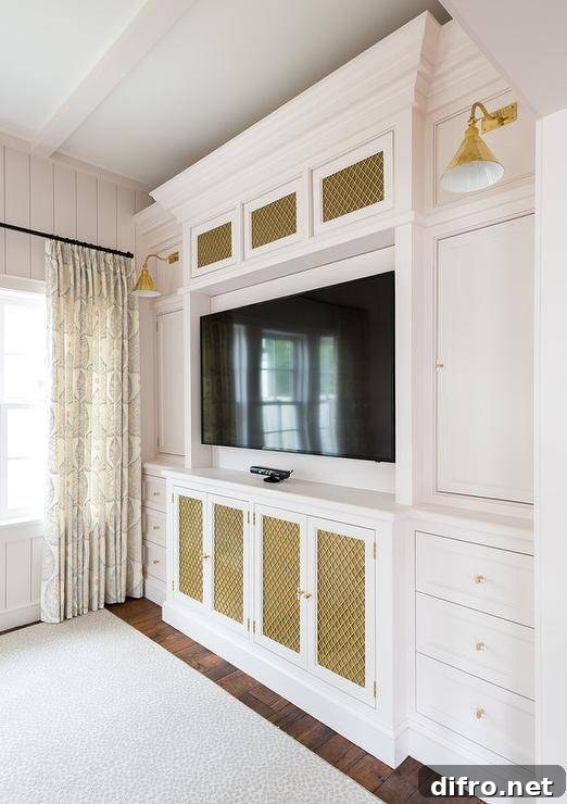 Brass metal grill front TV cabinets accented with brass and glass pulls flank a flat panel TV mounted in a niche between white cabinets and drawers lit by gold conical sconces.