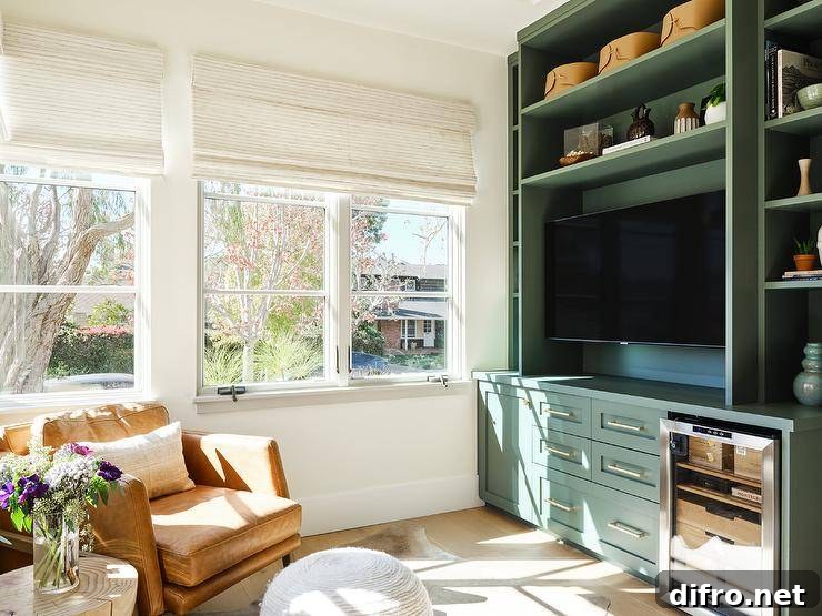 Family room features forest green TV built ins with a glass front mini fridge, brown leather chair and a white pouf as ottoman.