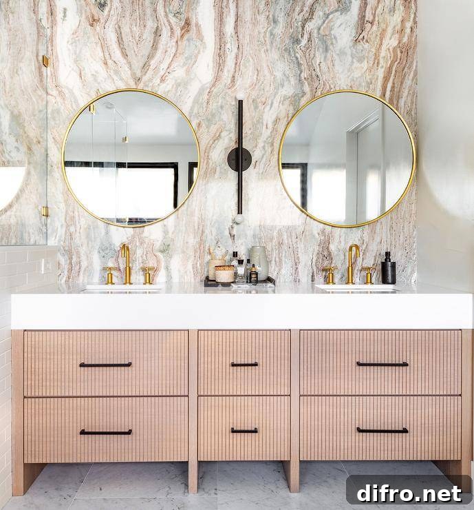 Modern bathroom features round brass mirrors mounted on a pink and gray marble slab accent wall over a beige reeded double washstand with brush gold faucets.