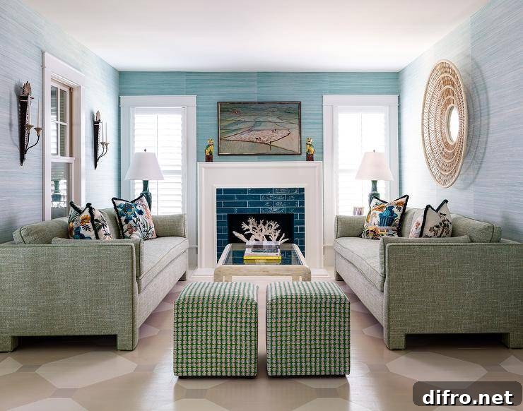 Glossy blue tiles are framed by a white fireplace mantel fixed beneath an art piece hung from an accent wall covered in blue grasscloth wallpaper. The fireplace is flanked by windows covered in white plantation shutters. A large round rattan mirror hangs from a wall clad in light blue grasscloth wallpaper over a green sofa placed on a tan geometric painted floor. The sofa faces a matching green sofa and is complemented with green cube stools.