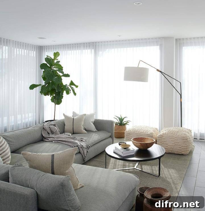 Framed by windows covered in sheer white curtains, this wonderfully designed living room features a fiddle leaf fig placed behind a pebble gray sectional complemented iwth French burlap pillows. The sofa is matched with a round black coffee table placed on a gray rug, while a black swing arm floor lamp with a white drum shade lights two white poufs.