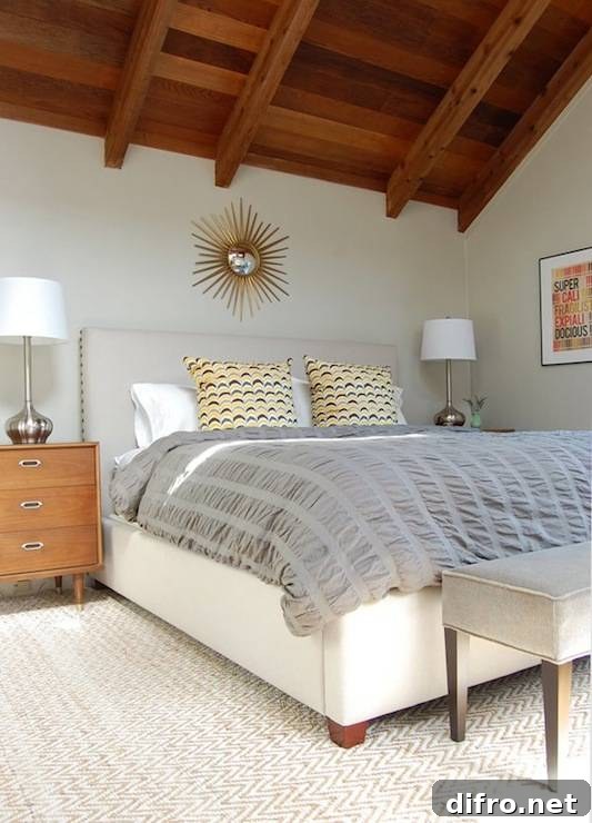 Attic bedroom with gold sunburst mirror over upholstered bed with gray pintuck comforter and retro yellow pillows.