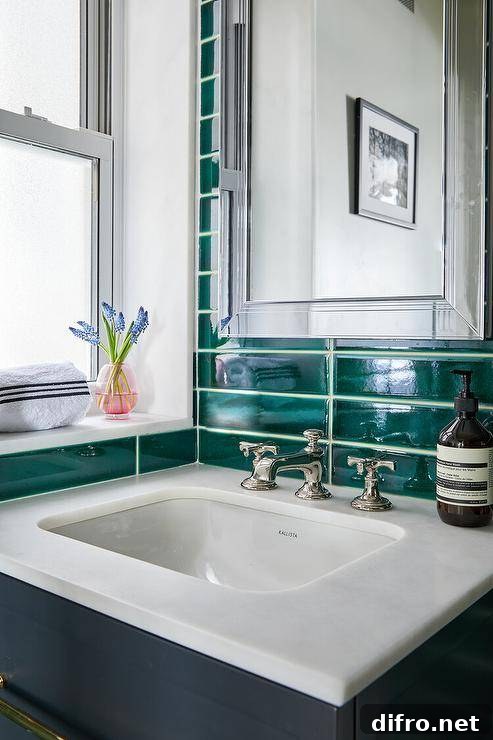 A blue floating washtsand with a polished nickel cross handle faucet is mounted against stacked emerald green tiles under a framed inset medicine cabinet.