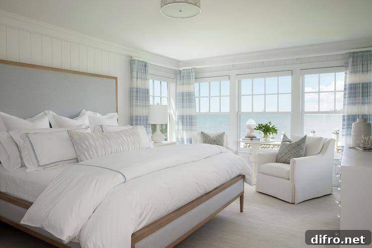 Brown and blue linen bed with white hotel bedding accented with blue borders in a cottage ocean view master bedroom. White linen skirted accent chairs join a white side table at the bedroom windows boasting a stunning view of a serene ocean and clear skies.