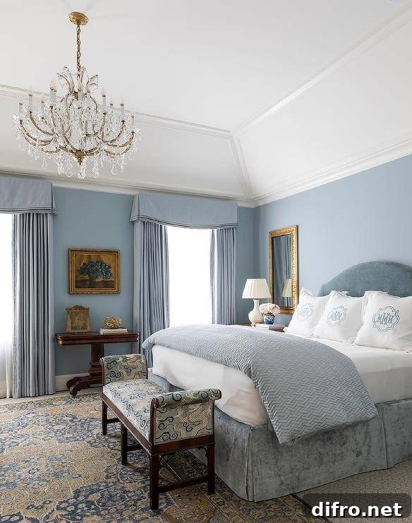 Sophisticated blue master bedroom features a blue velvet headboard that supports a bed dressed in a blue velvet pleated skirt, and white and blue monogrammed bedding, atop layered bedroom rugs.