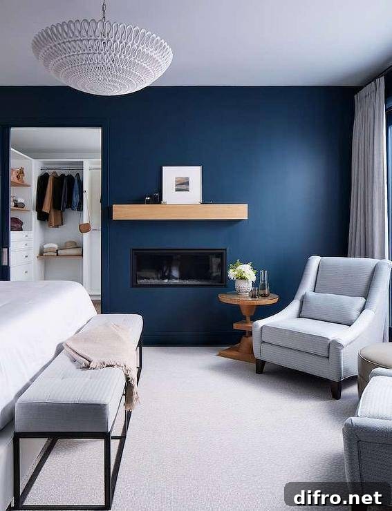 A modern inset fireplace with a styled chunky mantel is positioned in a blue painted bedroom wall beside a glass top round wooden accent table matched with a gray striped upholstered chair.