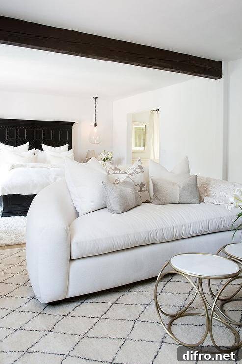Under a dark stained wood ceiling beam, a white blush curved sofa sits on a Moroccan style wood rug in a bedroom sitting area featuring a white and gold accent table.
