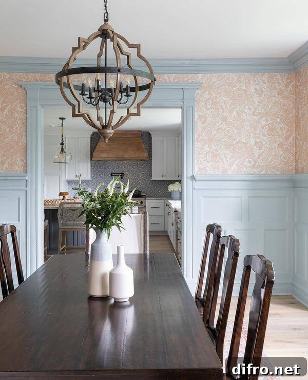 A wood and metal lantern hangs over a brown wood dining table surrounded by vintage wooden dining chairs. Walls half-covered in beige wallpaper are lined with blue crown moldings and finished with blue millwork.