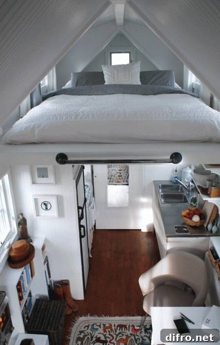 A small bed with white and grey bedding positioned above the living space that includes a small kitchen, modern chair, white desk, and bookshelf.